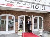 En kvinde i forretningstøj står ved den åbne dør til Dronninglund Hotel og byder gæsterne velkommen. Indgangen har et rødt tæppe og hvide planter med hotelskiltet over.