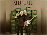 To smilende mænd står foran stablede grønne THY-ølkasser. Den ene holder trommestikker, den anden en guitar. Teksten ovenover lyder: MD-DUO Det Spiller Bår.