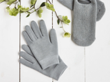 Grå strikhandsker og sokker på en hvid træoverflade med grønne grene og blomster i nærheden.