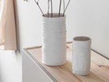 To hvide, strukturerede keramiske vaser står på en lys træoverflade. Den ene vase er høj og har små kviste indeni, mens den anden er lavere og tom. Baggrunden er minimalistisk og blødt oplyst.