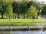En gruppe mennesker med golfbags står på en frodig grøn golfbane, omkranset af træer, med en dam og et springvand i forgrunden.