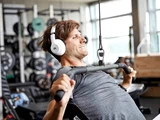 En mand med hvide hovedtelefoner og grå skjorte laver en lat pulldown-øvelse i et fitnesscenter med vægte og udstyr i baggrunden.