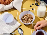 En morgenmadsscene med en skål granola, blåbær, en croissant, en flaske mælk, en kop te, en ske og en del af en avis på et gult kakkelbord.