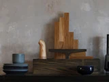 Et minimalistisk arrangement af køkkenredskaber af træ, mørkt service og skåle på en mørk overflade foran en grå væg med struktur.
