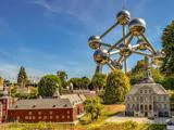 Atomium-monumentet tårner sig op bag miniaturebygninger og flag i en park på en solskinsdag, omgivet af grønt og blå himmel.