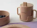 Beige spisesæt til småbørn med LOVE trykt på en drikkedunk og en skål, begge med hank, ved siden af en tallerken med mærket MINI og en skål med blåbær på en lilla måtte.