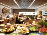 Moderne cafeteria med mennesker, der spiser ved borde; en buffet i forgrunden indeholder brød, frugt, ost og salat; varm belysning og moderne indretning.