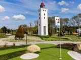 En minigolfbane med grønt kunstgræs, klipper og en hvid fyrtårnskopi under en blå himmel med spredte skyer. Et flag markeret med 12 står nær hullet.