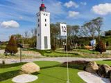 En minigolfbane med en hvid fyrtårnsstruktur, grønt kunstgræs, et flag med tallet 12, klipper og træer under en blå himmel med skyer.