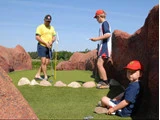 Tre personer er på en minigolfbane; en voksen spiller, et barn står med et scorekort, og et andet barn sidder på kunstgræsset ved siden af klipperne på en solrig dag.