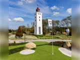 En minigolfbane med kunstgræs, klipper og et flag mærket med tallet 12, beliggende tæt på et højt, hvidt fyrtårn under en blå himmel med spredte skyer.