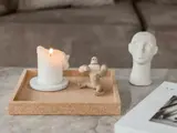 Et tændt hvidt stearinlys, en lille gorillafigur i træ og et hvidt skulpturelt hoved hviler på en korkbakke på et marmorbord ved siden af et magasin.