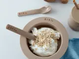 En beige skål mærket MINI fyldt med yoghurt og havregryn med en matchende ske mærket LOVE. En beige ske, en kop med sugerør og en blå klud ligger i nærheden på en hvid flade.