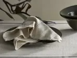 En beige stofserviet med tynde striber er pænt placeret på en mørk keramiktallerken, der står på en lys dug ved siden af en matchende skål.