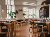 En moderne restaurant med træborde og sorte stole, dækket med tomme vinglas, omgivet af store vinduer og grønne potteplanter.