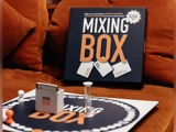 Et brætspil ved navn Mixing Box vises på en brun sofa med brikker, kort og spilleplade synlige i forgrunden.
