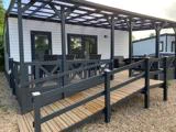 Et moderne mobilhome med en overdækket veranda, udendørs bord og stole samt en trærampe, der sikrer adgang til det grusbelagte område.