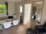 Moderne, kompakt opholdsrum med et spisebord og hvide stole, en lænestol, et stort vægspejl samt udsigt ind til et soveværelse og et badeværelse; vindue med brune gardiner.