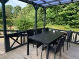 Et sort udendørs spisebord med seks stole står på en træterrasse under en pergola, omgivet af grønne træer og buske.