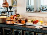 En morgenmadsbuffet med syltetøjsglas, ost, frisk frugt, grøntsager, kager og brød på en disk med et spejl og en spiseplads i baggrunden.