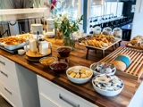 Morgenmadsbuffet med wienerbrød, brød, marmelade, smør, kornflakes i krukker, tallerkener og bestik på en trædisk i et moderne spiseområde.