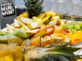Nærbillede af en farverig frugtplade med skiver af ananas, melon, druer, æbler og papaya. I baggrunden ved siden af frugten ses et skilt med teksten »Total Win-Win!«.
