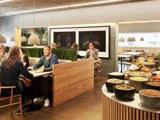 Folk sidder og taler ved træborde i et moderne cafeteria. I forgrunden står skåle med snacks eller nødder på en rund disk.