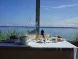 Et morgenbord med te, juice, kager og glas har udsigt til en rolig sø gennem et stort vindue på en solrig dag.