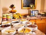 En morgenmadsbuffet med skåle med morgenmadsprodukter, tørret frugt og nødder, stablede farverige kopper, bananer, frisk frugt og juicekartoner på en træbordplade.