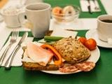 En morgenmadstallerken med brød med kerner, ost i skiver, skinke, salami, en skive rød peberfrugt, agurk og cherrytomater. Kaffe og redskaber står på bordet.