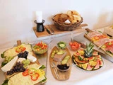 Et buffetbord med forskellige oste, vindruer, kød i skiver, friske grøntsager, brød i en kurv, vandmelonskiver og et stearinlys i en holder.