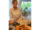 En smilende kvinde står indendørs bag et bord med croissanter, frugt og skåle med toppings, med glas og et lyst vindue i baggrunden.