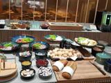 Et buffetbord med forskelligt brød, rundstykker, pålæg, oste, frisk frugt, pålæg og grøntsager, anrettet i skåle og på tallerkener, med redskaber og tallerkener i nærheden.