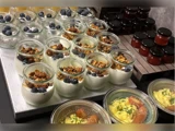 En udstilling af glas fyldt med yoghurt, granola og blåbær ved siden af glas med rød marmelade og beholdere med en gul skål med krydderurter.