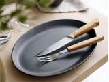 En sort oval tallerken med en gaffel og en savtakket kniv, begge med træhåndtag, placeret på et lyst træbord.