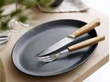 En sort oval tallerken med en gaffel og en savtakket kniv, begge med træhåndtag, placeret på et lyst træbord.