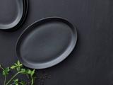 En sort oval tallerken på en mørk overflade med persilleblade og sorte peberkorn i nærheden; stablede tallerkener er delvist synlige i hjørnet.
