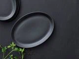 En sort oval tallerken på en mørk overflade med persilleblade og sorte peberkorn i nærheden; stablede tallerkener er delvist synlige i hjørnet.