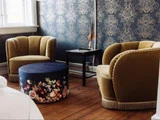To gule fløjlslænestole og en blomstermønstret rund ottoman står på et trægulv mod et blåblomstret tapet i et hyggeligt rum i vintagestil.