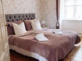 En pænt redt seng med lyserødt quiltet betræk, hvide puder og foldede håndklæder i et lyst soveværelse med blomstret tapet og et vindue, der lukker naturligt lys ind.