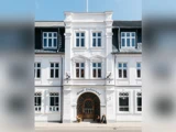 Billedet viser facaden på en hvid hotelbygning i tre etager med en central buet indgang med navnet Hotel Lidenlund og flere vinduer på hver etage.