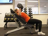 En person i orange skjorte laver incline dumbbell press på en bænk i et fitnesscenter med kettlebells og håndvægte i baggrunden.