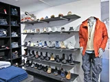 Tøjbutik med hylder fyldt med forskellige sko, sammenfoldede jeans til venstre og en udstillingsdukke iført en orange jakke og blå cargobukser til højre.