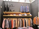 Tøjbutik med jakker, skjorter og bukser på bøjler, sammenfoldede varer på hylderne og udstillingsdukker klædt i jakkesæt under et ovenlys.