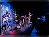 Et band optræder på scenen med guitarer og mikrofoner, mens publikum ser på. På baggrunden står der delvist »SMOKING«. Scenen er oplyst af blå og lilla lys.