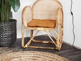 En rattan-stol med en rustfarvet pude står på et mørkt trægulv ved siden af et vævet tæppe og en stor potteplante i en sort kurv.