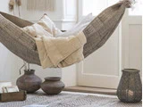 En mønstret hængekøje med beige puder og et tæppe hænger indendørs over et tæppe ved siden af lerkrukker, en lanterne og en stak bøger i et hyggeligt, lyst rum.