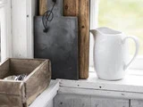 En hvid keramikkande står i vindueskarmen ved siden af en trækasse og to skærebrætter, et i træ og et i sort, i en rustik køkkenindretning.