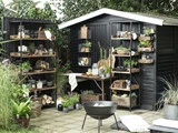 Et stilfuldt udekøkken med sorte hylder, trækasser, potteplanter, glas og køkkenting, der står op ad et sort skur omgivet af grønt og et bålsted foran.