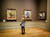 En kvinde med langt blondt hår står i et kunstgalleri og betragter tre indrammede malerier af blomster og natur, som hænger på væggen.